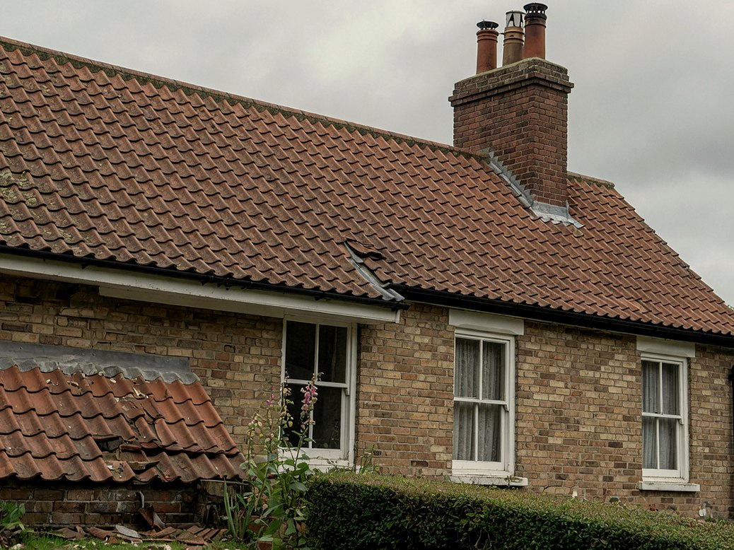 Brick house with a clay-tiled pitched roof, a missing tile, and a collapsed outbuilding roof in the foreground